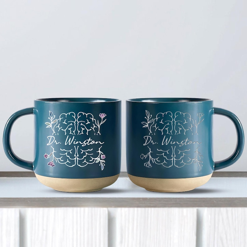 May include: Two teal ceramic mugs with a light tan base. Each mug features a white brain illustration with floral accents and the text "Dr. Winston". The mugs have a curved handle.