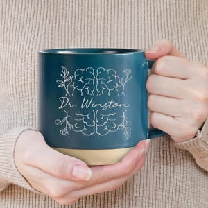 May include: A teal ceramic mug with a cream-colored base. The mug features a white brain illustration with floral accents and the text "Dr. Winston". The mug is held by a person.