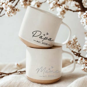 May include: Two stacked white ceramic mugs with a beige base. The top mug says "Papa - est. 2026" in black script, and the bottom mug says "Mama - est. 2026" in silver script. Both mugs have a heart symbol.