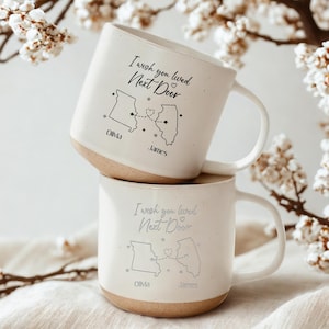 May include: Two stacked white ceramic mugs with a tan base. Each mug has the text "I wish you lived Next Door" and a map outline with names "Olivia" and "James". The mugs are decorated with a heart and star details.