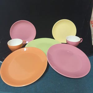 Puede incluir: Una colección de vajilla de cerámica colorida, que incluye platos y tazas de té. Los platos son en tonos rosa, amarillo, naranja y verde. Las tazas de té son naranjas y rosas. La vajilla está dispuesta sobre una superficie oscura.
