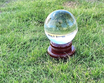 May include: A clear crystal ball sits atop a dark wooden stand, reflecting the surrounding green grass and sky. The sphere is a focal point, adding a touch of mystique to the outdoor setting. The base is a rich, dark brown.