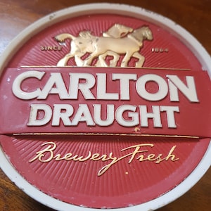 Puede incluir: Un posavasos redondo de color rojo y blanco con un diseño dorado. Presenta dos caballos tirando de un carro, el texto "Carlton Draught", y las palabras "Since 1864" y "Brewery Fresh".