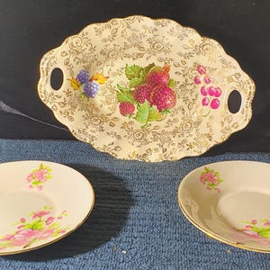 May include: Three vintage ceramic plates with floral designs. The center plate is an oval serving dish with a raised rim and handles. The dish features a floral pattern with a central design of fruit, including strawberries, raspberries, and grapes.