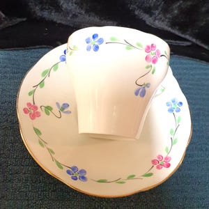 Może przedstawiać: Biały kubek do herbaty i spodek z pozłacanym rantem. Kubek i spodek są ozdobione delikatnym wzorem kwiatowym w odcieniach niebieskiego, różowego i zielonego.