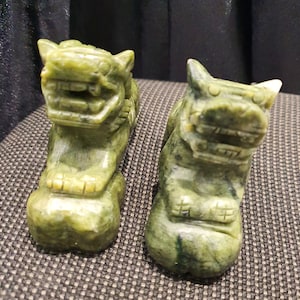 May include: Two green jade stone sculptures of foo dogs, traditional Chinese guardian lions, with intricate details and a textured finish.