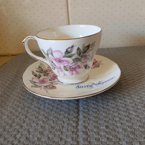 Könnte beinhalten: Weißes Tee-Set mit einem Blumendesign in Rosa und Grün. Tasse und Untertasse haben einen goldenen Rand. Auf der Untertasse steht in blauer Schrift "Silver Wedding".
