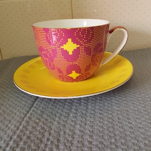 May include: A pink and yellow teacup and saucer set with a geometric pattern. The cup has a gold rim and the saucer has a gold rim and a gold band around the edge.