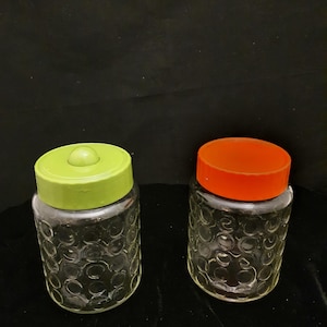 May include: Two glass jars with textured surfaces and coloured lids. The jar on the left has a lime green lid and the jar on the right has an orange lid.