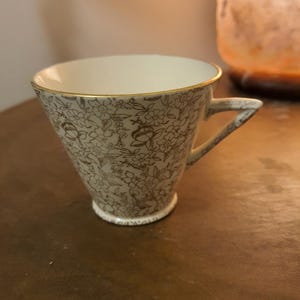 Puede incluir: Una taza de té blanca con un borde dorado y un patrón floral marrón. La taza tiene una forma acampanada y un asa.