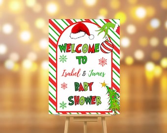 May include: A festive baby shower sign on an easel. The sign features a red and green striped border and the words "WELCOME TO Isabel & James BABY SHOWER" in green and red. Christmas-themed graphics include a Santa hat, Grinch's hand, and a Christmas tree.