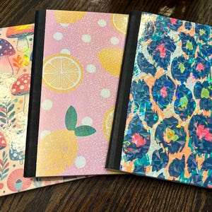 May include: Three notebooks with decorative covers. One features mushrooms, another lemons, and the third a leopard print. The covers have a holographic, glittery finish and black spines. The notebooks are arranged on a dark wood surface.