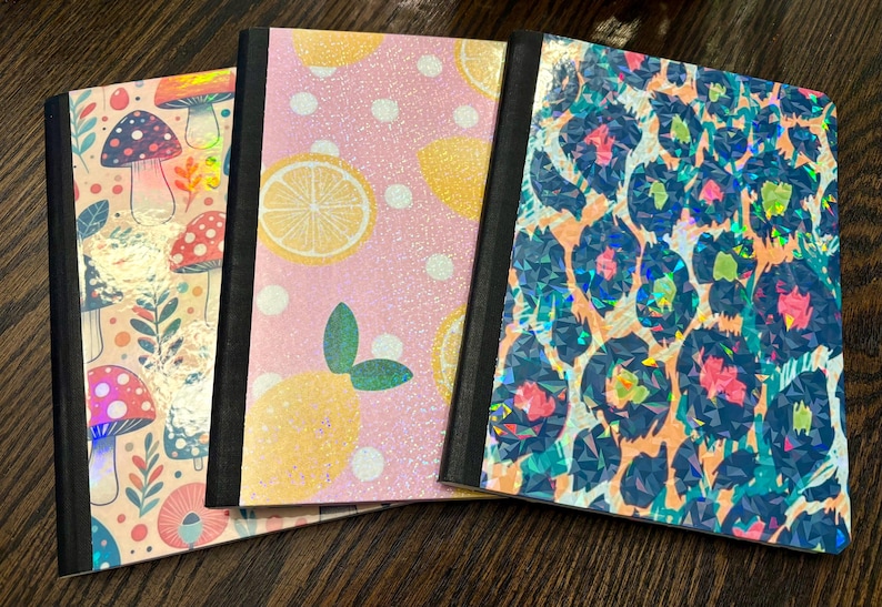 Pu&ograve; includere: Tre quaderni con copertine decorative. Uno presenta funghi, un altro limoni e il terzo una stampa leopardata. Le copertine hanno una finitura olografica e glitterata e dorsi neri. I quaderni sono disposti su una superficie di legno scuro.
