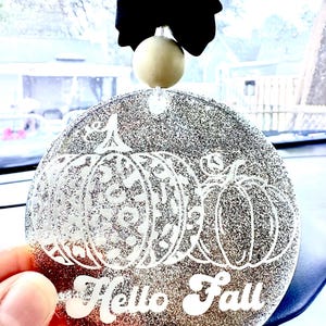 May include: A round, silver glitter ornament featuring a white graphic of two pumpkins with leopard print patterns and the text "Hello Fall" in a script font. The ornament is topped with a natural wooden bead and a black fabric bow.