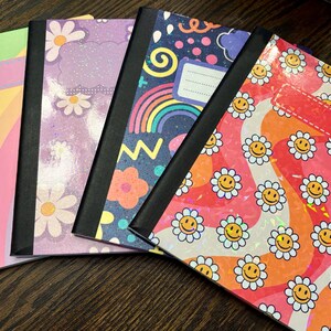 Pu&ograve; includere: Quattro quaderni con copertine colorate con arcobaleni, margherite e faccine sorridenti. Ogni quaderno ha un dorso nero e un'etichetta vuota sul davanti.