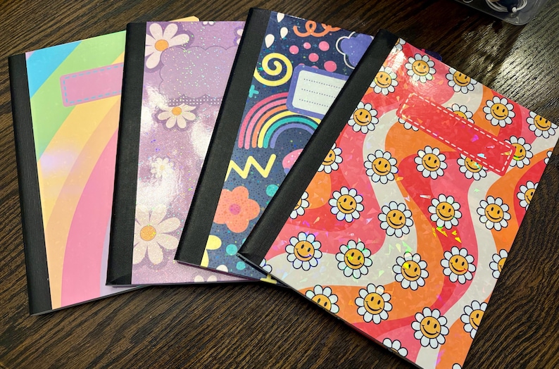 Pu&ograve; includere: Quattro quaderni con copertine colorate con arcobaleni, margherite e faccine sorridenti. Ogni quaderno ha un dorso nero e un'etichetta vuota sul davanti.