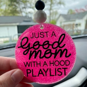 May include: A pink glittery car charm with the text "JUST A good mom WITH A HOOD PLAYLIST" in black script. The charm is round and has a string with black and white beads for hanging.