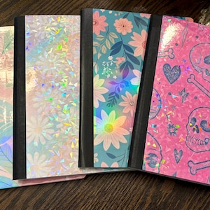 Pu&ograve; includere: Una collezione di cinque quaderni con vari design. Le copertine presentano motivi floreali, texture olografiche e un motivo a teschio rosa. Ogni quaderno ha un dorso nero e un design di copertina unico, offrendo una variet&agrave; di stili.