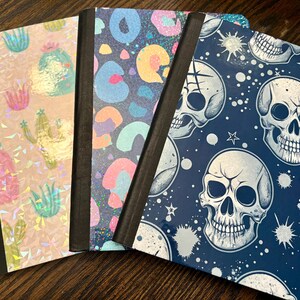Pu&ograve; includere: Quattro quaderni con copertine uniche. Uno presenta cactus olografici, un altro una stampa leopardata colorata e un terzo teschi su sfondo blu navy. I quaderni hanno dorsi neri e sono disposti su una superficie di legno.