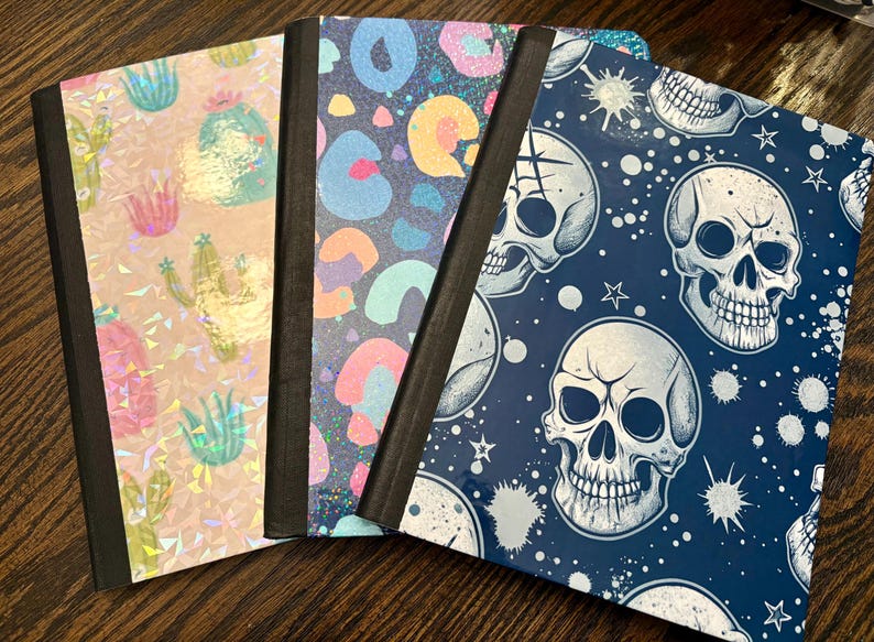 Pu&ograve; includere: Quattro quaderni con copertine uniche. Uno presenta cactus olografici, un altro una stampa leopardata colorata e un terzo teschi su sfondo blu navy. I quaderni hanno dorsi neri e sono disposti su una superficie di legno.