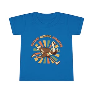 Op de afbeelding: Een blauw T-shirt met een cartoon struisvogel die rent. De struisvogel is bruin en wit, met een groen-witte streep. De tekst rond de struisvogel luidt "FUTURE OLYMPIC SPRINTER" in oranje en geel.
