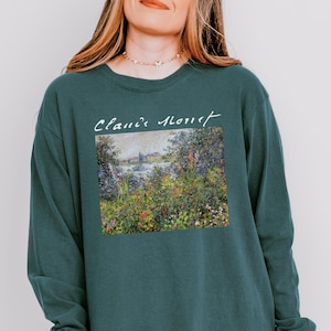 May include: Teal long-sleeve sweatshirt featuring a Claude Monet painting print. The artwork depicts a colorful landscape with a body of water and buildings. The text "Claude Monet" is above the artwork in a cursive font. The words "Blue Spruce" are at the bottom left.