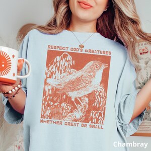 May include: Pale blue t-shirt featuring a rust-coloured illustration of a bird on a branch, with the words "Respect God's Creatures Whether Great or Small." The shirt is labelled "Chambray" at the bottom.