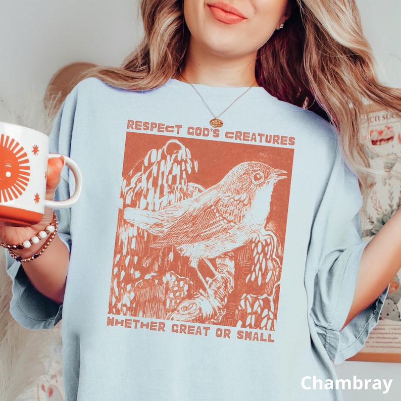May include: Pale blue t-shirt featuring a rust-coloured illustration of a bird on a branch, with the words "Respect God's Creatures Whether Great or Small." The shirt is labelled "Chambray" at the bottom.
