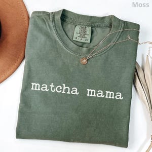 May include: A moss green short-sleeved t-shirt with a crew neckline and the text "matcha mama" printed in white across the chest. The shirt features a Comfort Colors brand label inside the collar and is paired with a delicate gold-toned necklace.