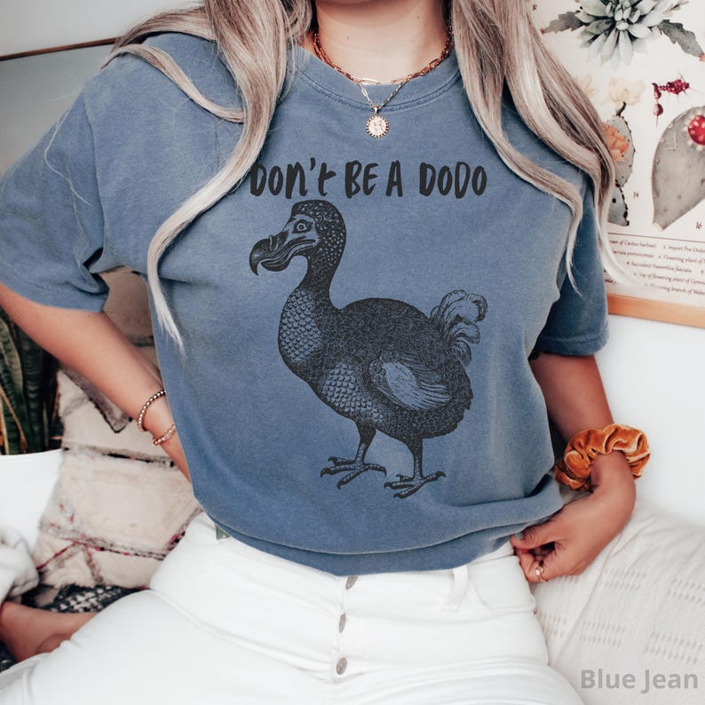 May include: Blue jean-colored t-shirt with a black graphic of a dodo bird and the text "DON'T BE A DODO" in black. The shirt is a short-sleeved, crew neck style.