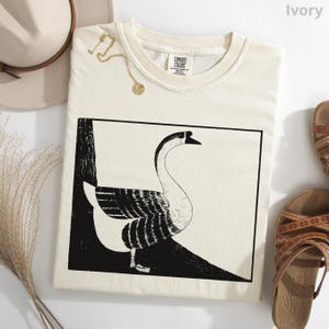 May include: Ivory-colored Comfort Colors t-shirt featuring a black and white illustration of a goose within a rectangular frame. A gold necklace and a beige hat are placed near the shirt. Brown sandals are also visible.
