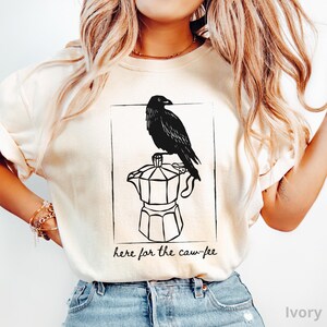 May include: Ivory-colored t-shirt featuring a black illustration of a raven perched atop a coffee pot. The design is enclosed in a rectangular frame, with the phrase "here for the caw-fee" printed below in a handwritten style.