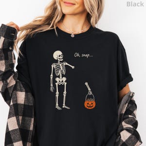 May include: A black t-shirt featuring a graphic of a skeleton with one detached arm holding an orange jack-o'-lantern candy bucket. The text "Oh, snap..." is printed in white above the skeleton. The shirt is worn with a black and grey plaid flannel shirt.