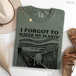 Peut inclure: T-shirt vert mousse avec l'inscription "I FORGOT TO WATER MY PLANTS!" et un graphisme inspir&eacute; de "Le Cri" d'Edvard Munch. Un collier dor&eacute;, un chapeau beige et des sandales marron sont &eacute;galement visibles.