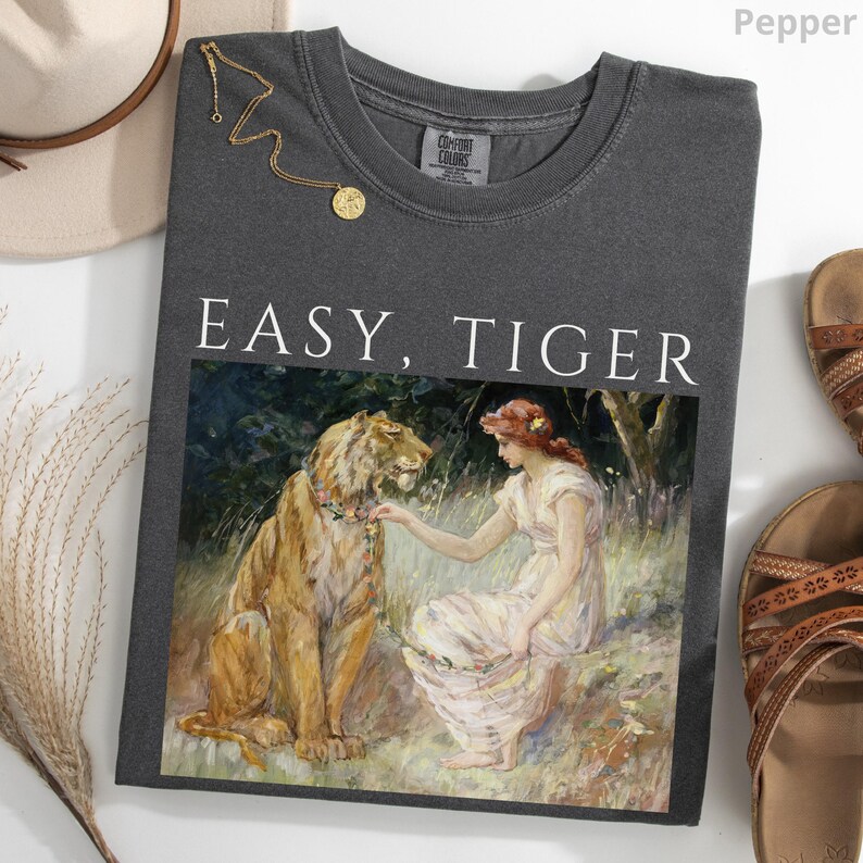 May include: Pepper-colored Comfort Colors t-shirt with the text "EASY, TIGER" above a painting of a tiger and a woman. A gold necklace and sandals are also visible.