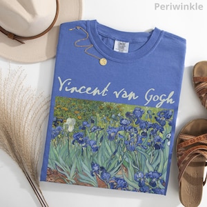 May include: A periwinkle blue t-shirt featuring the text Vincent van Gogh above a printed graphic of irises. The shirt is displayed alongside a beige hat, a gold chain necklace, and brown sandals, with the word Periwinkle visible in the corner.