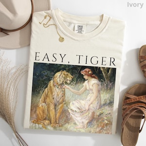 May include: Ivory-colored sweatshirt with the words "EASY, TIGER" printed in black. The graphic depicts a woman and a tiger in a forest setting. A gold necklace and a hat are visible. The sweatshirt is paired with sandals.