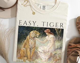 May include: Ivory-colored sweatshirt with the words "EASY, TIGER" printed in black. The graphic depicts a woman and a tiger in a forest setting. A gold necklace and a hat are visible. The sweatshirt is paired with sandals.