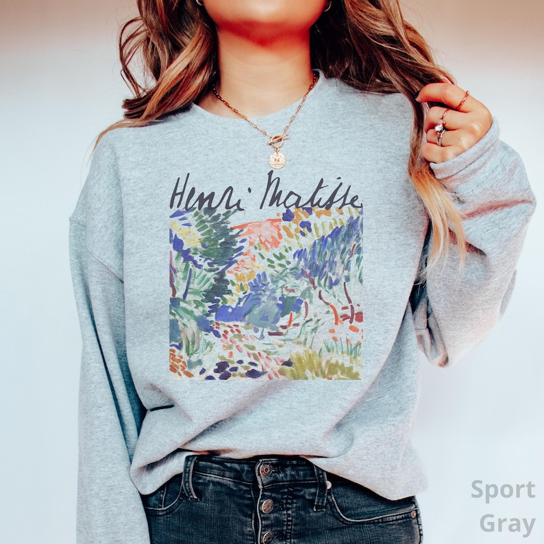 Puede incluir: Sudadera gris deportiva con una colorida pintura de paisaje de Henri Matisse. La obra de arte incluye tonos verdes, azules, naranjas y rosas. El nombre del artista est&aacute; escrito encima de la obra en escritura negra.
