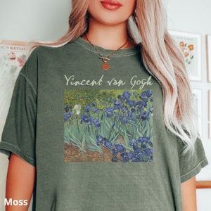 May include: A moss green short-sleeve t-shirt featuring a graphic print of Vincent van Gogh's Irises painting. The text Vincent van Gogh is printed in a light colour above the artwork, which depicts blue flowers and green foliage.