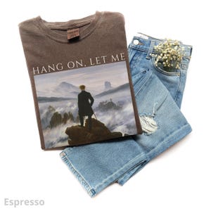 May include: A brown t-shirt with the text "Hang on, let me" printed on the front. The shirt features a black and white image of a man standing on a mountaintop with fog in the background.