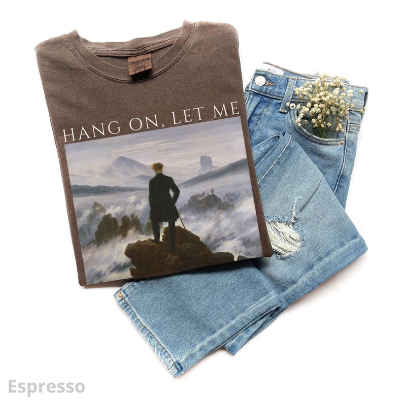 May include: A brown t-shirt with the text "Hang on, let me" printed on the front. The shirt features a black and white image of a man standing on a mountaintop with fog in the background.