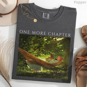 May include: Pepper-colored t-shirt with the text "ONE MORE CHAPTER" above a painting of a person reading in a hammock. A gold necklace and sandals are next to the shirt.