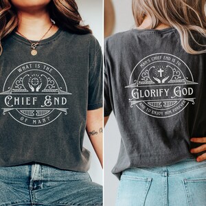 May include: A dark gray t-shirt with a white graphic design on the front and back. The front design says "What is the Chief End of Man?" with a cross and hands in a circle. The back design says "Man's Chief End is to Glorify God and to Enjoy Him Forever" with a cross and hands in a circle.