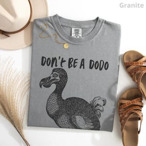 May include: A gray t-shirt with the text "DON'T BE A DODO" and a black illustration of a dodo bird. The shirt is accompanied by a gold necklace, a beige hat, and brown sandals.