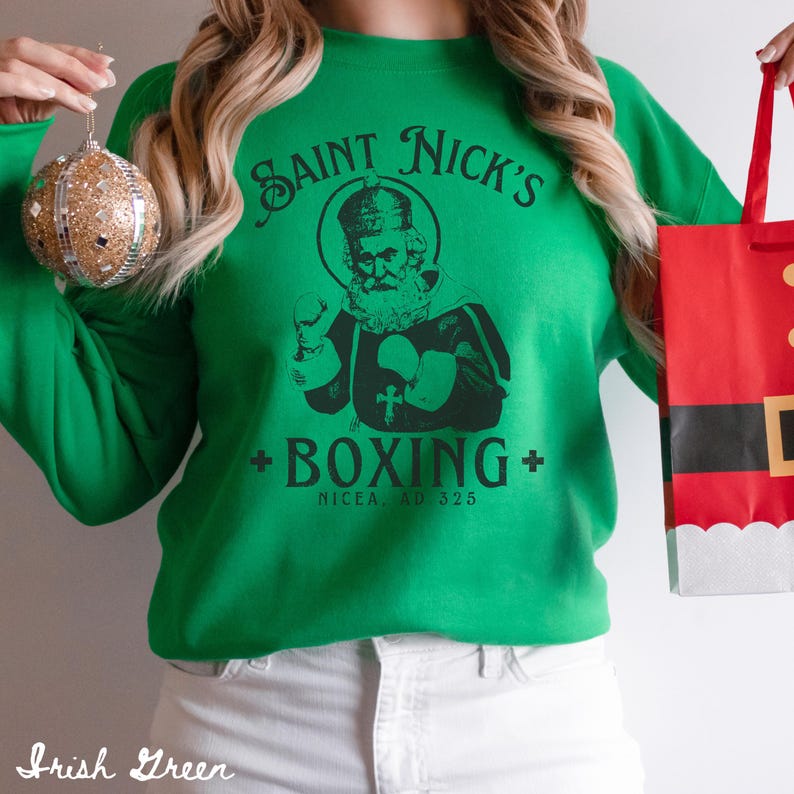 Op de afbeelding: Groen sweatshirt met een zwarte afbeelding van Sinterklaas die boksen. De afbeelding bevat de tekst "Saint Nick's Boxing" en "Nicaea, AD 325".