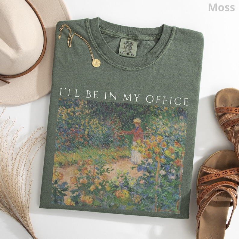 May include: A moss green t-shirt with the text "I'll Be In My Office" above a colorful impressionist painting. A gold necklace, a beige hat, and brown sandals are arranged around the shirt.