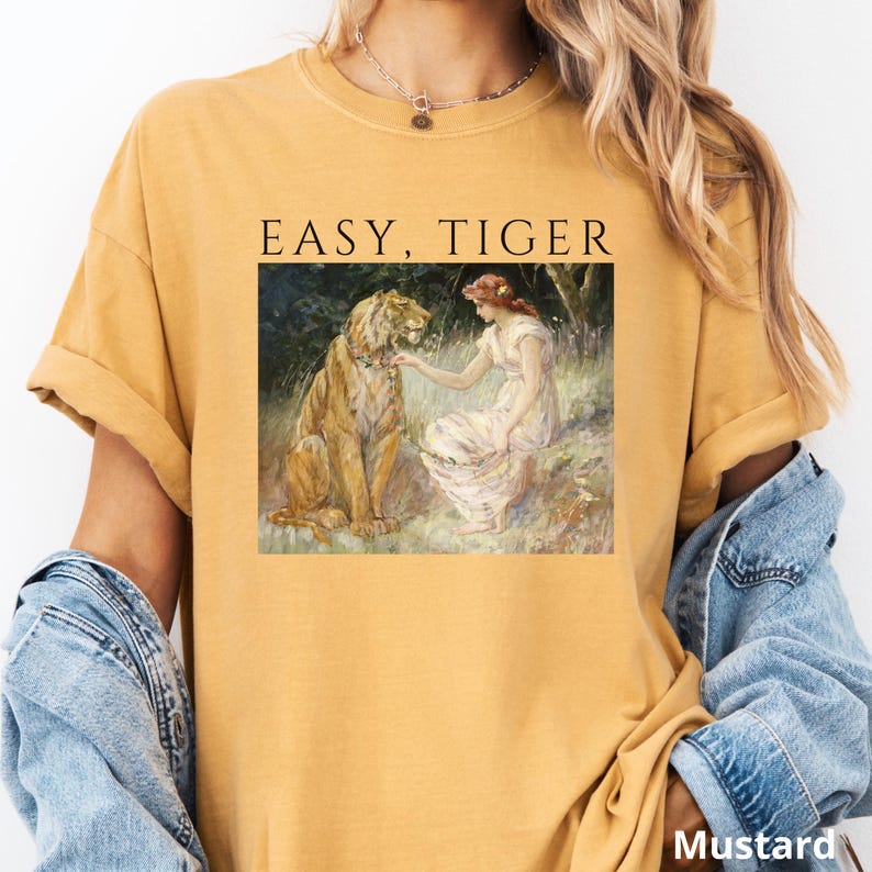May include: Mustard-colored t-shirt featuring a vintage-style illustration of a lion and a woman with the text "EASY, TIGER" above the image. The shirt has rolled-up sleeves and a relaxed fit.