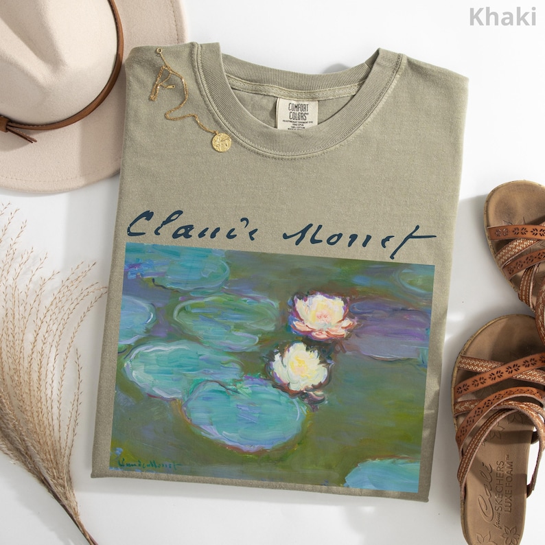 May include: Khaki-colored t-shirt featuring a print of Claude Monet's "Water Lilies" painting. The shirt has a crew neck and short sleeves. A gold necklace and a beige hat are next to the shirt. Brown sandals are also in the image.