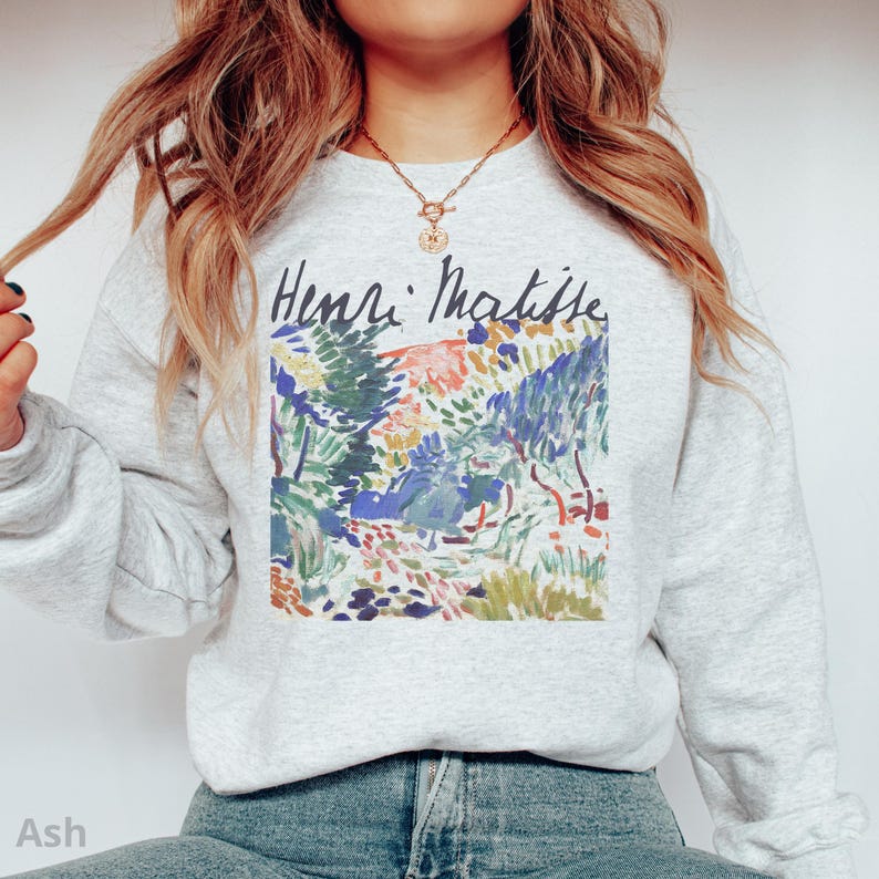 Puede incluir: Sudadera gris ceniza con una impresi&oacute;n colorida de una pintura de paisaje con el texto "Henri Matisse" en escritura negra. La obra de arte incluye tonos verdes, azules, amarillos y rojos. La sudadera es de material suave.